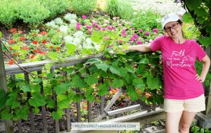 White woman standing in a garden, wearing a pink shirt that says: Faith can move Mountains