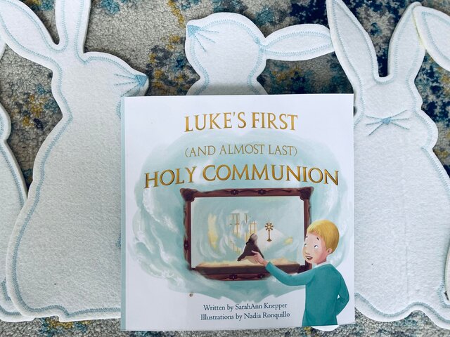 Photo of a paperback book set on a background of white bunnies. Book title is: "Luke's First (And Almost Last) Holy Communion"