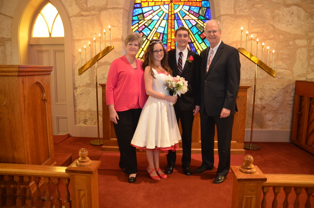 Image of a white family after a wedding, at the front of church. From left to right: bride's mother in pink, bride in short white dress with pink accessories, groom in black suit, bride's father in black suit.